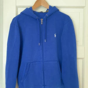 Blå hoodie från Polo Ralph Lauren - Blå hoodie från Polo Ralph Lauren med dragkedja framtill, snörning i huvan och klassisk broderad logga på bröstet. Tröjan har känguruficka och är perfekt för en chill och stilren look. Priset går att diskutera vid snabb affär.