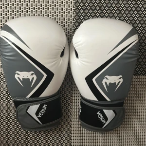 Venum boxningshandskar 12oz svart/vit - Säljer ett par snygga boxningshandskar från Venum i svart, vitt och grått. Handskarna har coolt ormmotiv och tydlig Venum-logga på ovansidan. De är tillverkade i syntetmaterial och har kardborreknäppning för extra stöd runt handleden. Använda fåtal gånger. Funkar till sparring och säck.