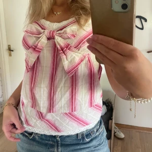 Mönstrad linnetopp med rosett - Säljer en söt linnetopp med tunna axelband och stor rosett framtill. Toppen är vit med rosa ränder och har spetsdetalj vid halsringningen. Materialet är lätt och luftigt, perfekt för varma dagar. 