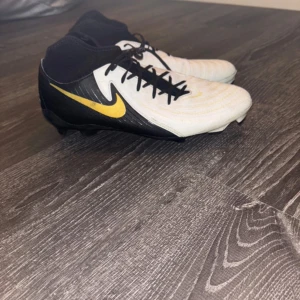 Nike Fotbollsskor Phantom Luna II Pro svart/vit/gul  - Säljer ett par Nike fotbollsskor i svart och vitt med snygga gula detaljer och klassisk swoosh på sidan. Skorna har snörning, högre strumpliknande skaft och mönstrad ovandel för extra grepp. Perfekta för dig som vill sticka ut på planen. (knappt använda
