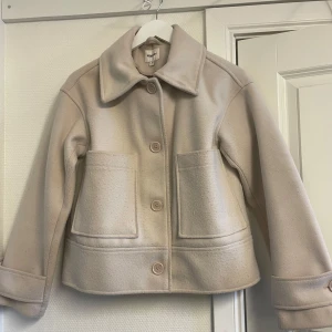 Beige ulljacka från Kappahl - Stilren beige jacka från Kappahl i ullmix med stora fickor framtill och klassisk krage. Jackan har stora knappar och raka ärmar, perfekt för dig som gillar en clean och enkel look. Passformen är normal och materialet känns mjukt och varmt. Jackan är något nopprig vilket tydliggörs på sista bilden☺️