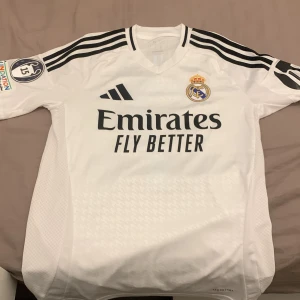 Real Madrid Vini Jr. 7 matchtröja XS - Säljer en officiell Real Madrid fotbollströja från Adidas i vitt med svarta detaljer och Vini Jr. 7 på ryggen. Tröjan har korta ärmar, klubbmärke på bröstet, broderad Adidas-logga och tryckt sponsor. Tillverkad i lätt och ventilerande material. !! TAR BARA KONTANT SÅ SKRIV OM DU VILL KÖPA KAN MÖTAS MELLAN MÄRSTA - NORRVIKEN