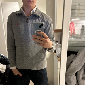 Ralph Lauren half zip tröja  - Grå långärmad tröja från Polo Ralph Lauren med klassisk röd logga på bröstet. Tröjan har en snygg half zip-dragkedja med läderdetalj och hög krage. Tillverkad i mjuk bomull och passar perfekt till lager-på-lager-stil. Hör av dig vid ytterligare frågor🤗