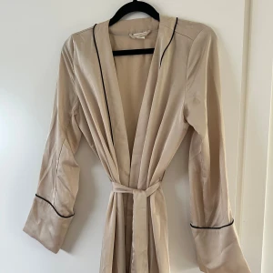 Morgonrock i satin  - Säljer en beige morgonrock från H&M i satinliknande material. Den har långa ärmar, svarta kantdetaljer och knytskärp i midjan. 