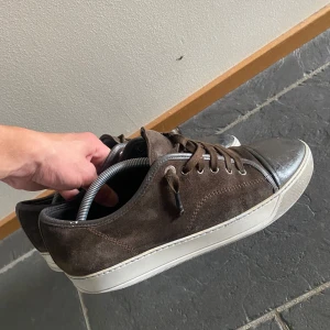 lanvin - Säljer ett par bruna sneakers från Lanvin med ovandel i mocka och tåparti i mörkbrunt skinn. Skorna har vita gummisulor och klassisk snörning. Perfekta för dig som gillar stilrena och lyxiga sneakers med en twist. Först till kvarn inne sulan är dock ej så bra men inte så dåligt att det är hål 