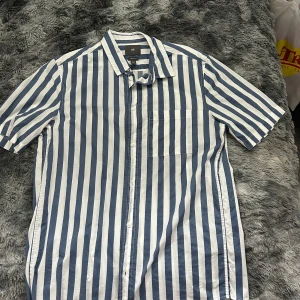 Randig kortärmad skjorta från H&M - Snygg kortärmad skjorta från H&M i regular fit med breda vertikala blå och vita ränder. Skjortan har klassisk krage, bröstficka och knäppning framtill. Perfekt för en avslappnad och stilren look.
