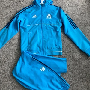 Marseille tracksuit - Säljer en blå marseille dress från Adidas med Olympique de Marseille-logga och vita detaljer som är ganska svår att få tag i. Tröjan har huva, dragkedja framtill och svarta ränder längs ärmarna. Perfekt för fotboll eller träning, tillverkad i polyester och med lång ärm.