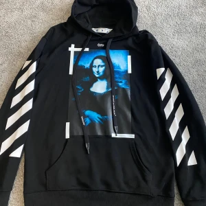 Off-White Mona Lisa hoodie svart/blå - Svart hoodie från Off-White med ikoniskt Mona Lisa-tryck i blått på bröstet och vita pilar på ryggen. Ärmarna har vita diagonala ränder och luvan har snörning. Perfekt statement-plagg för dig som gillar streetwear och konst. Passar även S