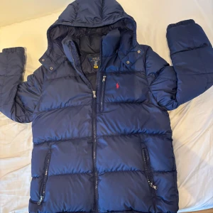 Ralph lauren Jacka  - Snygg marinblå pufferjacka från Polo Ralph Lauren i storlek XL (barn storlek). Jackan är köpt förra vintern. Extremt nöjd med jackan men måste Tyvär göra mig av me den på grund utav att den e för liten. Jackan har en avtagbar luva och så att man kan knäppa runt halsen villket är otroligt tacksamt när de blåser. Jackan har en inne ficka perfekt för förvaring av väder saker samt en ficka på bröstet och två på sidan. Defekter, jackan har ett litet hål på baksidan och en dragkedja som har fastnat.⛷️❄️