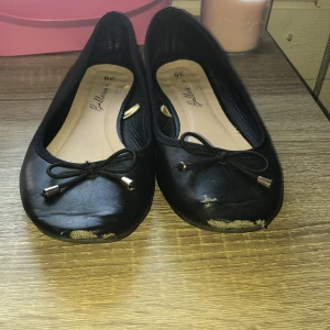 Svarta ballerinaskor med rosett, strl 38 - Svarta ballerinaskor i syntetmaterial med rund tå och dekorativ rosett framtill med guldfärgade detaljer. Platt sula och klassisk design som passar till många outfits. Enkla att ta på och av, perfekta för dig som gillar stilrena skor.