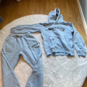 Ljusblå hoodie från Hoodrich - Säljer en ljusblå hoodie från Hoodrich med svart tryck framtill. Tröjan har huva, magficka och ribbade muddar vid ärmslut och nederkant. Perfekt för chill dagar och streetwear-stil.