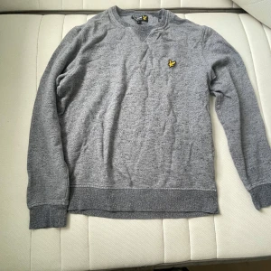 Grå tröja från Lyle & Scott, storlek L - Grå långärmad tröja från Lyle & Scott med klassisk rund hals och gul logga på bröstet. Tröjan har ribbade muddar och är tillverkad i mjuk bomullsmix, perfekt för en chill och stilren look.