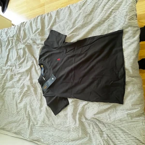 Svart t-shirt från Polo Ralph Lauren - Svart t-shirt från Polo Ralph Lauren med klassisk rund hals och korta ärmar. T-shirten har en liten röd broderad logga på bröstet och är gjord i mjuk bomull. Perfekt för en clean och stilren look.