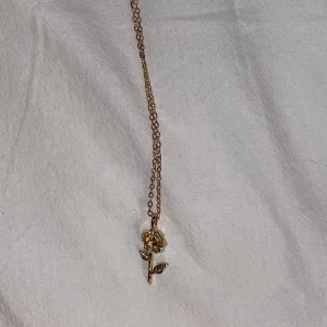 Guldigt halsband med blomma - Snyggt halsband i guldton med ett hängsmycke format som en blomma. Kedjan är tunn och smidig, och blomman har detaljerade blad och kronblad som ger en cool och trendig vibe. Perfekt för dig som gillar blommiga smycken och vill ha något som sticker ut.
