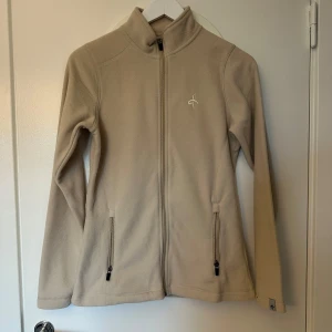 Beige fleecejacka från Cross Sportswear - Beige fleecejacka från Cross Sportswear med hel dragkedja framtill och två fickor med dragkedja. Jackan har hög krage och broderad logga på bröstet. Perfekt för kyliga dagar och enkel att matcha med andra plagg.