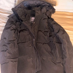 Svart wyndham Canada Goose jacka (black label) - Säljer en svart dunjacka från Canada Goose med pälskantad huva och klassisk logga på ärmen fast i svart (black label edition) Jackan har dragkedja och knappar framtill, samt flera praktiska fickor. Perfekt för kalla vinterdagar och riktigt varm. Även äkta päls top kvalite går även att dra bort pälsen och sätta på igen med dragkedjan. Har knappast varit andvänd bara ett par ggr men ser helt ny ut 