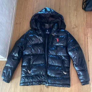 Svart pufferjacka från Polo Ralph Lauren - Snygg svart pufferjacka från Polo Ralph Lauren med glansig finish, huva och röda loggan broderad på bröstet. Jackan har dragkedjor på fickorna och framtill, samt quiltad design som ger en cool look. Perfekt för kalla dagar.