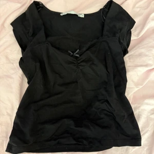 Svart topp med spets och rosett - Svart topp från lager 157 med v-ringning, spetskant och en liten söt rosett framtill. Toppen är ärmlös och har en figurnära passform, perfekt att styla med jeans eller kjol. Tillverkad i mjuk bomull för en bekväm känsla.