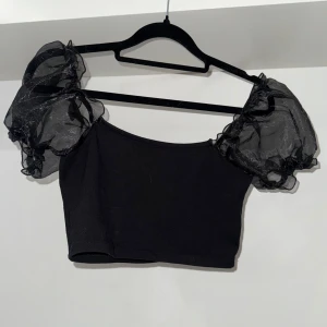 Svart croppad topp med puffärmar från Topshop - Säljer en svart croppad topp från Topshop med genomskinliga puffärmar. Toppen har en tight passform och ärmlös kropp med korta, voluminösa ärmar som ger en trendig look. Perfekt för dig som vill sticka ut med en cool och stilren vibe. Mycket fint skick!