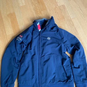 Blå vindjacka från Henri Lloyd M - Snygg marinblå vindjacka från Henri Lloyd med broderad logga på bröstet och brittisk flagga på ärmen. Jackan har dragkedja, ribbade muddar med vita ränder och meshfoder.