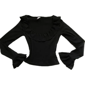 Svart volangtopp med långa ärmar - Säljer en svart topp med långa ärmar och volangdetaljer både vid halsringningen och ärmsluten. Toppen har en figurnära passform och är tillverkad i ett mjukt material som känns skönt mot huden. Perfekt för dig som gillar en stilren look med extra detaljer.