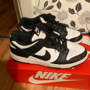 Nike Dunk Low svart/vit sneakers - Nike Dunk Low sneakers i klassisk svart och vit färg. Skorna har en låg siluett, svart swoosh och snörning framtill. Ovandelen är i skinn med perforerad tåbox för extra andningsförmåga. Perfekta för dig som gillar streetstyle och vill ha en ikonisk look.