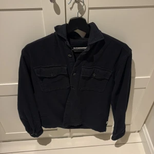 Svart overshirt från Jack & Jones - Stilren svart overshirt från Jack & Jones Premium med klassisk krage, två bröstfickor med knappar och långa ärmar. Skjortan är tillverkad i ett mjukt bomullstyg och har en avslappnad passform som funkar till många olika looks.