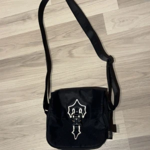 Trapstar svart axelväska med spänne - Svart axelväska från Trapstar med justerbar rem och coolt metallspänne framtill. Väskan har flera fack med dragkedja och detaljer i vitt på locket. Materialet är syntet och väskan har en urban vibe som passar till streetwear.