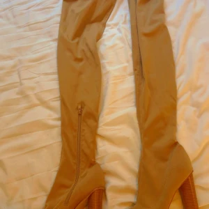 Beiga overknee stövlar med klack - Snygga beiga overknee stövlar med hög klack och dragkedja på insidan. Skorna har en rund tå och är tillverkade i ett mjukt syntetmaterial. Perfekta för att lyfta din outfit med en trendig touch.