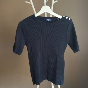 Marinblå ribbad topp från GANT med knappar - Marinblå  ribbad topp från GANT med korta ärmar och rund halsringning. Snygga silvriga knappar på ena axeln ger en extra detalj. Toppen är figurnära och tillverkad i ett stretchigt material som sitter skönt på kroppen.🌸