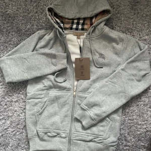 Grå burberry zip -  Snygg grå hoodie från Burberry med klassiskt rutigt foder i huvan, dragkedja framtill och broderad logga på bröstet. Mjuk insida och praktiska fickor. Perfekt för dig som vill ha en stilren och lyxig look. Helt ny och oanvänd med prislappen på, tveka inte att fråga frågor