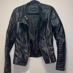 Svart skinnjacka från Missay XXS - Snygg svart skinnjacka från Missay i storlek XXS. Jackan har coola detaljer som silvriga dragkedjor, knappar vid kragen och quiltade partier på axlar och ärmar. Justerbara spännen i midjan och flera fickor framtill. Perfekt för en edgy look.