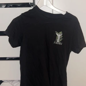 Svart Amiri t-shirt med ängeltryck - Svart t-shirt från Amiri med stort tryck av en vit ängel och gröna blad på ryggen samt mindre tryck på bröstet. Klassisk passform och rund halsringning. Perfekt statement-plagg för dig som gillar streetwear och coola prints.