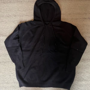 Helt ny Svart hoodie i merinoull - Svart hoodie i mjuk merinoull med klassisk huva och snörning. Tröjan har långa ärmar, ribbade muddar och en relaxed passform. Perfekt för dig som vill ha en stilren och bekväm look under kyliga dagar.
