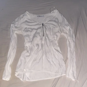 Vit långärmad topp med dragkedja - Vit långärmad topp från Gina Tricot med v-ringning och en snygg dragkedja framtill. Tunt och lätt material som passar perfekt till våren eller sommaren. Enkel och stilren design som är lätt att matcha.