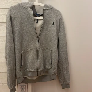 Grå hoodie från Polo Ralph Lauren - Grå hoodie från Polo Ralph Lauren med dragkedja, huva och två stora fickor framtill. Klassisk broderad logga på bröstet och ribbade muddar. Tyget har blivit väldigt nött på ena armen som ni ser på bilden 