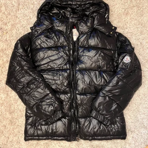 Svart Moncler dunjacka med huva - Säljer en svart glansig dunjacka från Moncler med huva och dragkedja. Jackan har klassisk pufferstil, logotyp på ärmen och flera praktiska fickor. Perfekt för kalla dagar och riktigt snygg streetstil.