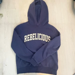 Blå hoodie REBELICIOUS Gina Tricot - Mörkblå hoodie från Gina Tricot med trycket REBELICIOUS i vitt framtill. Klassisk modell med huva och långa ärmar, ribbade muddar och mjukt material. Perfekt för dig som gillar en chill och avslappnad stil.