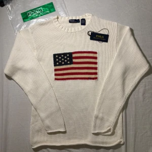 Vit stickad tröja från Polo Ralph Lauren - Vit stickad tröja från Polo Ralph Lauren med amerikansk flagga broderad på bröstet. Tröjan har rund halsringning, ribbade muddar och lång ärm. Klassisk och stilren design som sticker ut med flaggdetaljen.