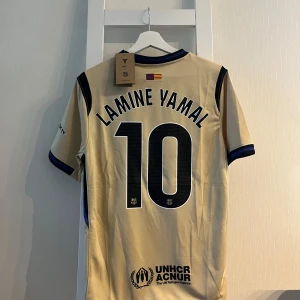 Beige FC Barcelona Lamine Yamal tröja - Säljer en beige FC Barcelona fotbollströja med Lamine Yamal och nummer 10 på ryggen. Tröjan har svarta detaljer, blå och svarta ränder vid krage och ärmslut, samt klubbmärke och sponsorloggor framtill. Tillverkad i lätt och ventilerande material.