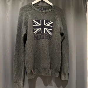 Grön stickad tröja från Morris - Snygg grön stickad tröja från Morris med Union Jack-flagga och broderad logga på bröstet. Tröjan har rund halsringning, ribbade muddar och lång ärm.