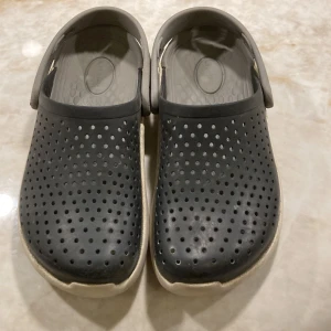 Svarta Crocs sandaler med vit sula - Svarta Crocs sandaler med perforerad ovandel och vit, tjock sula. Skorna har en justerbar hälrem och är tillverkade i syntetmaterial. Perfekta för chill dagar eller när du vill ha något bekvämt på fötterna.Storlek 31.