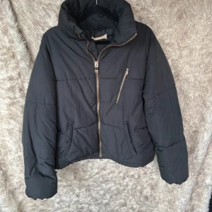 Svart pufferjacka med dragkedjor storlek 164 - Svart pufferjacka med hög krage och coola dragkedjedetaljer, inklusive sned bröstficka och två sidofickor. Jackan har en oversized vibe och är perfekt för kalla dagar. Tillverkad i syntetmaterial och har långa ärmar.