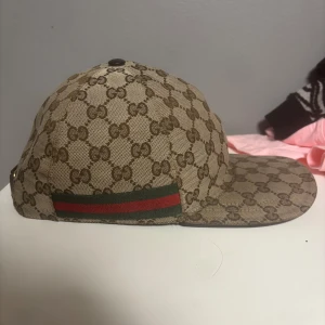 Gucci keps med GG-mönster, storlek L - Snygg keps från Gucci i beige och brunt med klassiskt GG-monogram över hela kepsen. Grön och röd rand på sidan och justerbar rem baktill i mörkbrunt skinn. Perfekt accessoar för dig som gillar lyxiga detaljer och streetstyle. Pris är diskuterbart 