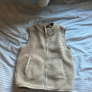 Beige fleeceväst från SOC - Mysig beige fleeceväst från SOC med dragkedja framtill och två fickor. Västen har en fluffig teddylook och är perfekt att slänga på sig över en hoodie eller tröja. Enkel och stilren design som funkar till många outfits.