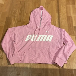 Rosa hoodie från Puma med logga - Säljer en rosa hoodie från Puma med stor vit logga framtill. Tröjan har huva med snörning och en klassisk känguruficka. Perfekt för dig som gillar sportig och avslappnad stil.