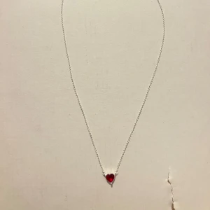 Halsband med röd hjärtberlock från Pandora - Superfint halsband i silver från Pandora med en liten röd hjärtformad berlock. Kedjan är tunn och elegant, perfekt för att ge en söt touch till din stil. Låset är klassiskt och har en liten märkbricka med Pandora-loggan.