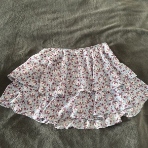 Blommig volangkjol från Zara - Supersöt kort volangkjol från Zara i ljusblått med blommigt mönster i rosa, lila och beige toner. Finns shorts under!! Kjolen har flera lager volanger som ger en flowy känsla och elastisk midja för extra komfort. Perfekt för dig som gillar romantisk och feminin stil.