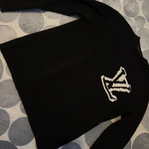 Svart stickad tröja Louis Vuitton - Svart stickad tröja från Louis Vuitton med stort vitt LV-logo på bröstet. Klassisk rund halsringning och långa ärmar. Perfekt för dig som vill ha en stilren och lyxig look med en tydlig logga.