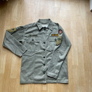 Grön overshirt från Ralph Lauren - Militärinspirerad grön overshirt från Ralph Lauren med coola patchar och broderade detaljer på bröstet och ärmarna. Jackan har två stora bröstfickor med knappar och klassisk krage. Perfekt för dig som gillar streetstyle och utility-vibe.